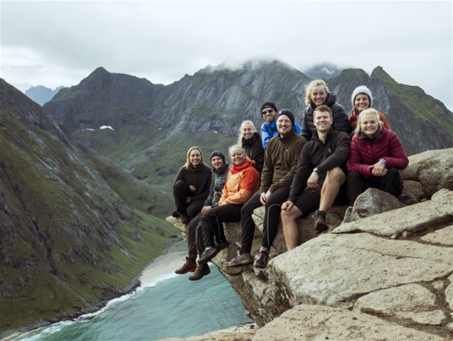 Traineer på Norgestur i flott, sitter på kanten av et fjell med fjell og innsjø i bakgrunnen